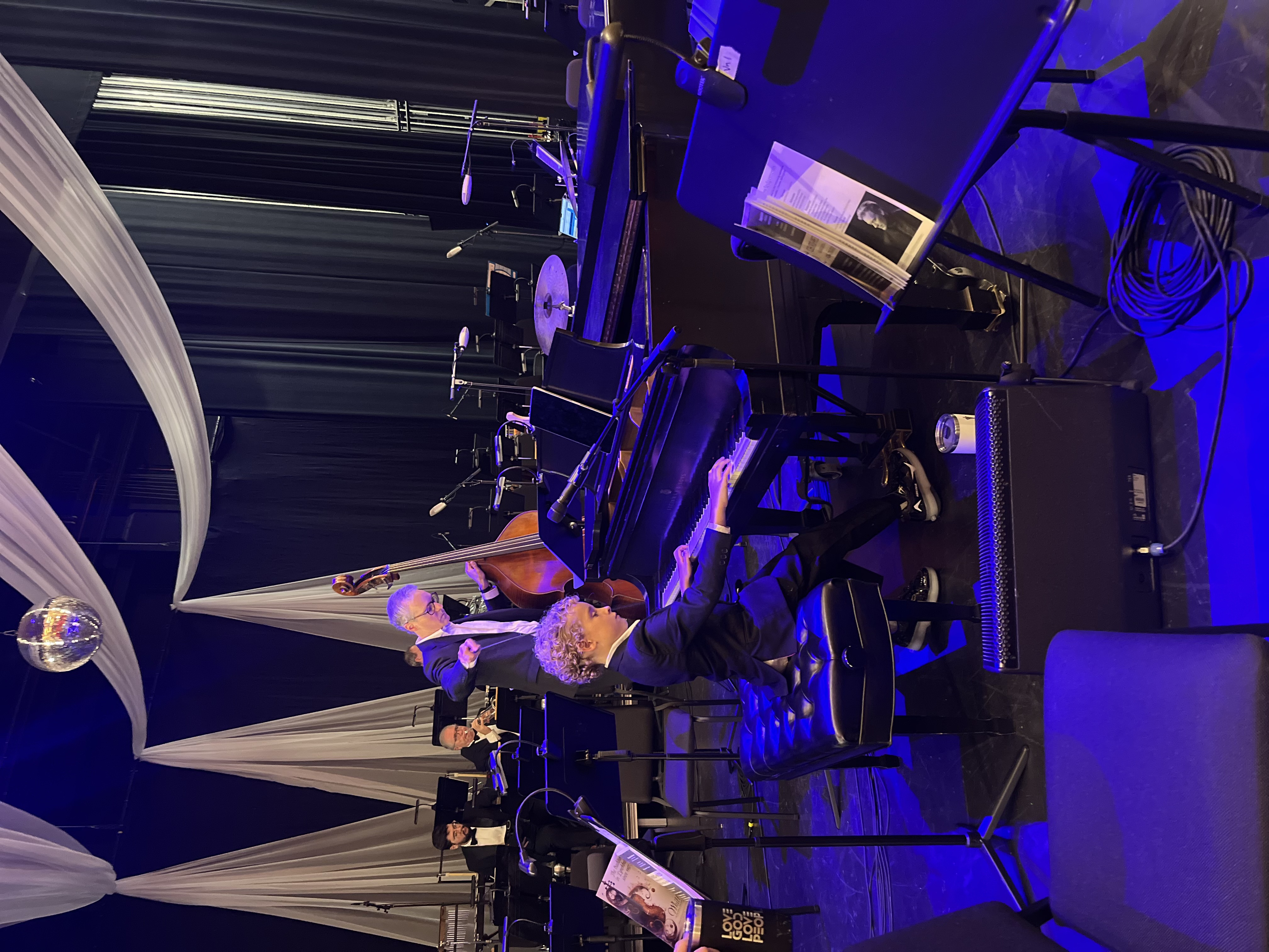 Leland at a Steinway grand piano on stage with orchestra musicians behind him